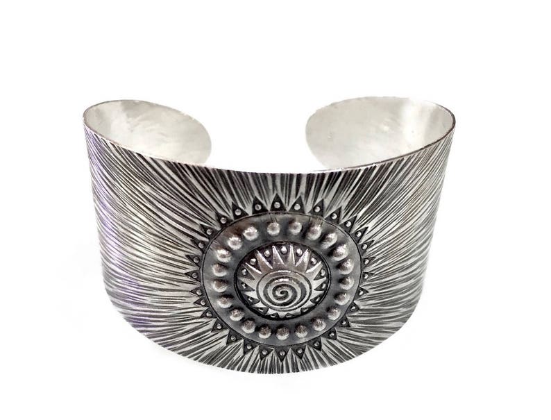May include: A wide, silver cuff bracelet with a sunburst design. The bracelet features a central circular medallion with a spiral and dot pattern. The cuff has a textured, radial pattern and is set against a white background.