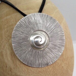 May include: A silver pendant with a spiral design in the centre and a textured, sunburst pattern around the edge. The pendant is hanging from a black cord.
