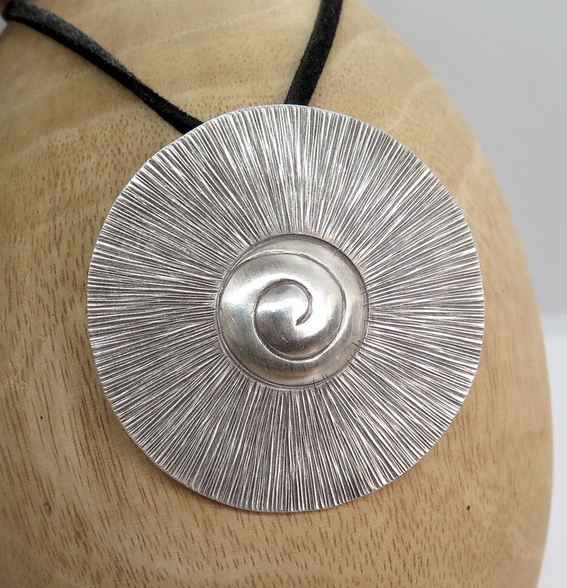 May include: A silver pendant with a spiral design in the centre and a textured, sunburst pattern around the edge. The pendant is hanging from a black cord.
