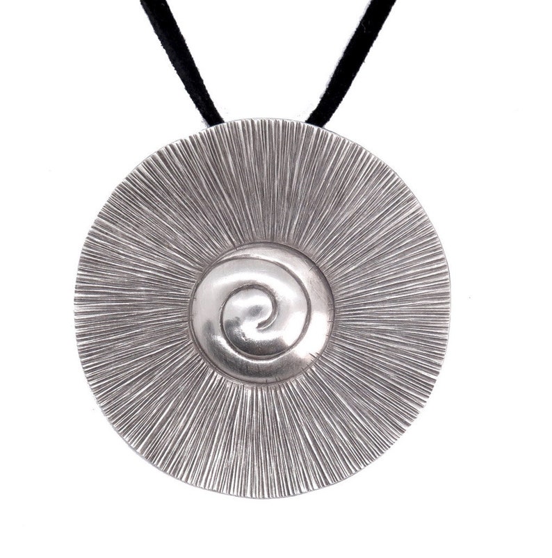 May include: A silver pendant necklace with a circular design featuring a spiral in the centre. The pendant is attached to a black cord.