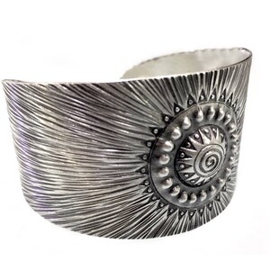 May include: A wide silver cuff bracelet with a sunburst design. The bracelet features a central circular motif with a spiral and is surrounded by small, raised dots and triangular shapes. The surface has a textured, radial pattern.