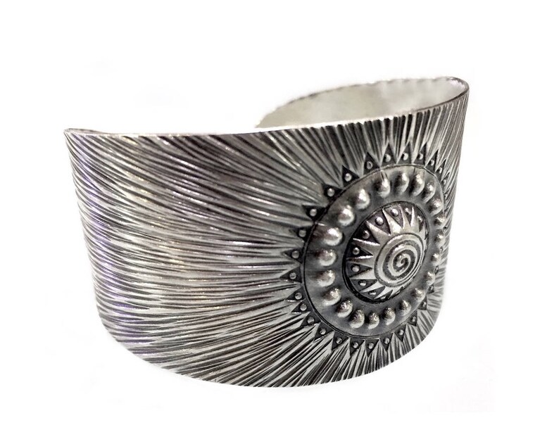 May include: A wide silver cuff bracelet with a sunburst design. The bracelet features a central circular motif with a spiral and is surrounded by small, raised dots and triangular shapes. The surface has a textured, radial pattern.