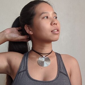 May include: A woman wearing a gray tank top with a silver circular pendant necklace. The pendant has a textured, concentric circle design.