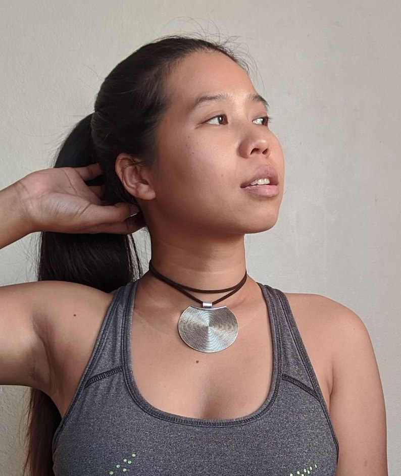 May include: A woman wearing a gray tank top with a silver circular pendant necklace. The pendant has a textured, concentric circle design.