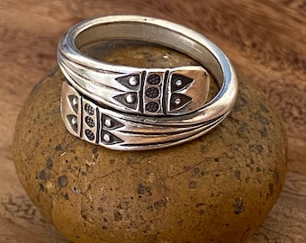 May include: A silver ring with a unique design featuring a repeating pattern of triangles and circles. The ring is made of two bands that are intertwined.