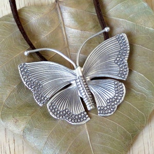 May include: A silver butterfly pendant necklace with a detailed textured design. The pendant is hanging on a brown leather cord.