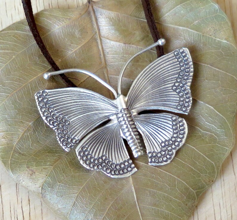 May include: A silver butterfly pendant necklace with a detailed textured design. The pendant is hanging on a brown leather cord.