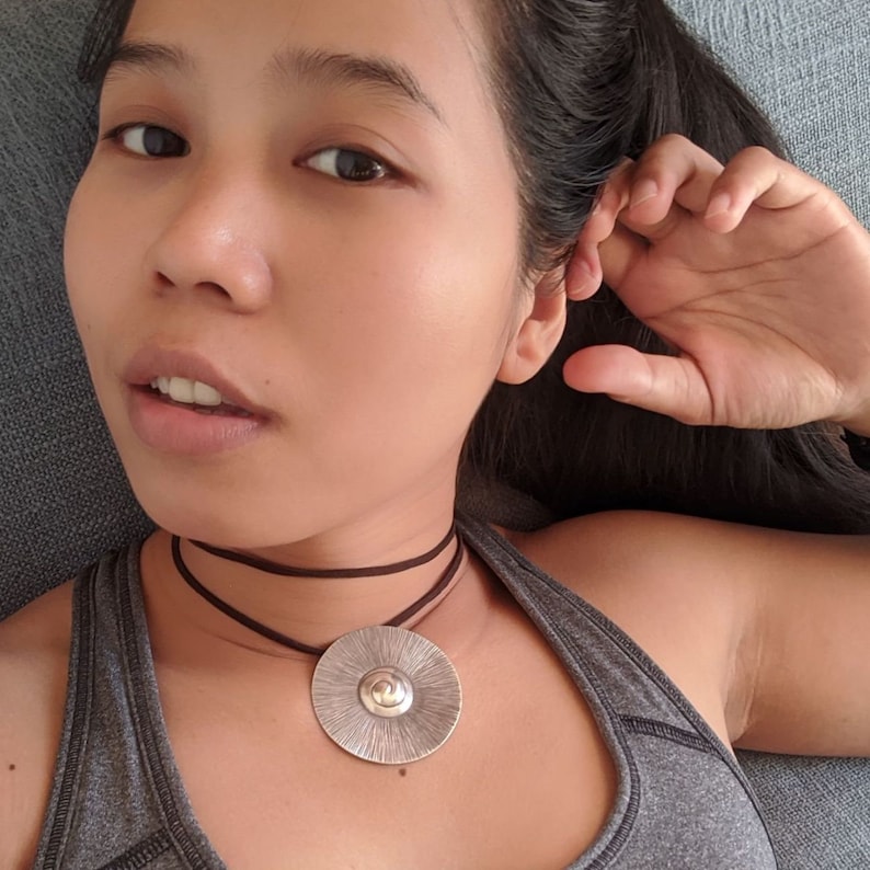 May include: A woman wearing a grey tank top with a brown leather choker necklace and a silver pendant with a textured circular design.