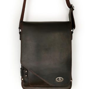 May include: A dark brown leather messenger bag with a shoulder strap. The bag has a rectangular shape with a flap closure and a metal clasp. The strap is also brown and appears to be adjustable. The bag is designed for carrying items.