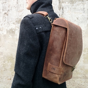 May include: A brown leather backpack with a flap closure and a metal buckle. The backpack has a small, rectangular patch with the text "mmm leather goods" on it.
