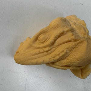 Op de afbeelding: Een gedetailleerde, lichtgele sculptuur van een reptielenkop, mogelijk een dinosaurus of draak. De sculptuur heeft een getextureerde huid, een prominent oog en een gedetailleerde snuit. De kop ligt op een wit oppervlak.