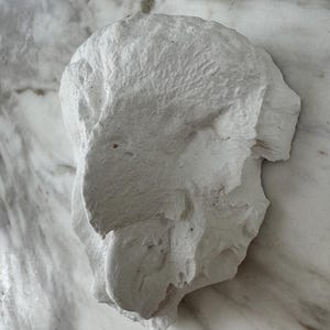 Op de afbeelding: Een ruw gevormde, gebroken witte sculptuur met een getextureerd oppervlak. De vorm lijkt een abstracte weergave van een hoofd of gezicht te zijn, met zichtbare contouren en een ruwe, onafgewerkte uitstraling. De achtergrond is gemarmerd.