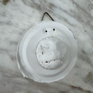 Op de afbeelding: Een witte, cirkelvormige wanddecoratie met een gebeeldhouwd gezicht. Het stuk heeft een verhoogd gezicht in een cirkelvormig frame, met een metalen draad om op te hangen. De textuur lijkt ruw en handgemaakt.