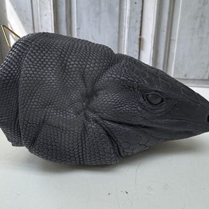 May include: A detailed, dark gray lizard head sculpture with a textured, scaled surface. The sculpture features a realistic eye and a gold-colored metal loop attached to the top, suggesting it may be a decorative item or a mask.
