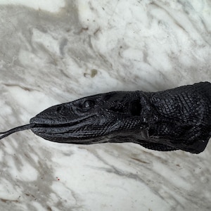 Könnte beinhalten: Eine detaillierte, schwarze Skulptur eines Reptilienkopfes mit einer langen, gespaltenen Zunge. Die Skulptur hat ein strukturiertes, schuppiges Aussehen und befindet sich vor einem marmorierten Hintergrund.