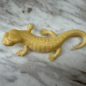 Op de afbeelding: Een lichtgele hagedissenfiguur met een getextureerd oppervlak. De hagedis heeft vier poten, een lange staart en een gedetailleerde kop. Het beeldje ligt op een marmerpatroon.