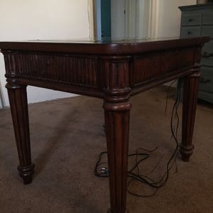 Op de afbeelding: Een donkerbruine houten bijzettafel met een glazen blad en gedraaide poten. De tafel staat op een bruin tapijt.