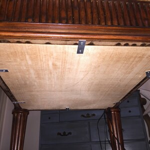 Op de afbeelding: De onderkant van een houten tafel met een bevestigd multiplexpaneel. Het paneel is bevestigd met metalen beugels. De tafel heeft een donkerbruine afwerking.
