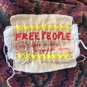 Puede incluir: Primer plano de una etiqueta de ropa con las palabras "FREE PEOPLE" bordadas en rojo. La etiqueta es de color crema con detalles de punto de cruz amarillo y el texto "made in china / fabriqu&eacute; en chine". La tela detr&aacute;s de la etiqueta tiene un patr&oacute;n oscuro.