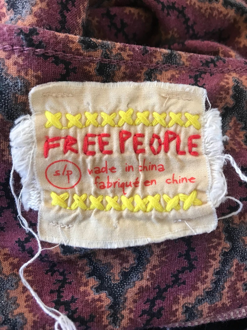Puede incluir: Primer plano de una etiqueta de ropa con las palabras "FREE PEOPLE" bordadas en rojo. La etiqueta es de color crema con detalles de punto de cruz amarillo y el texto "made in china / fabriqu&eacute; en chine". La tela detr&aacute;s de la etiqueta tiene un patr&oacute;n oscuro.