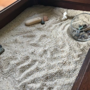 Op de afbeelding: Een houten dienblad met een zandtuin. Het zand is wit en heeft een paar kleine stenen en een kleine witte steen in de vorm van een vogel. Er is een kleine glazen kom met gekleurde stenen.