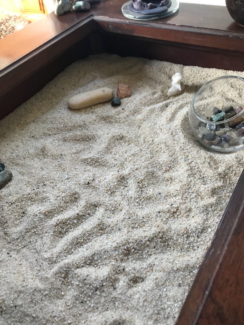 Op de afbeelding: Een houten dienblad met een zandtuin. Het zand is wit en heeft een paar kleine stenen en een kleine witte steen in de vorm van een vogel. Er is een kleine glazen kom met gekleurde stenen.