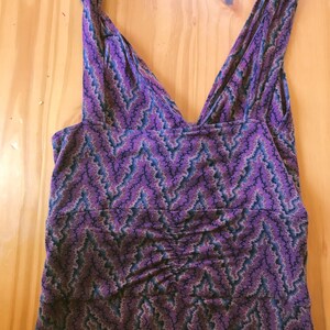 Puede incluir: Vestido morado sin mangas con cuello en V y estampado de zigzag en tonos morado, azul y beige. El vestido tiene un detalle fruncido en la cintura y est&aacute; hecho de una tela suave y fluida. El vestido est&aacute; plano sobre una superficie de madera.