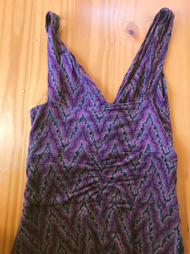 Puede incluir: Vestido morado sin mangas con cuello en V y estampado de zigzag en tonos morado, azul y beige. El vestido tiene un detalle fruncido en la cintura y est&aacute; hecho de una tela suave y fluida. El vestido est&aacute; plano sobre una superficie de madera.