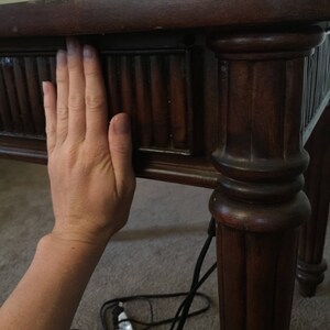 Op de afbeelding: Close-up van een donkerbruine houten tafel met een hand die op de bovenkant rust. De tafel heeft een decoratieve, gegroefde rand en een gedraaid poot.