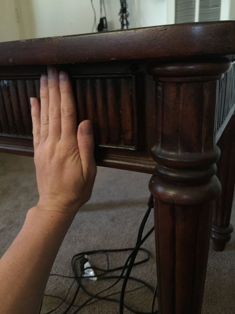 Op de afbeelding: Close-up van een donkerbruine houten tafel met een hand die op de bovenkant rust. De tafel heeft een decoratieve, gegroefde rand en een gedraaid poot.