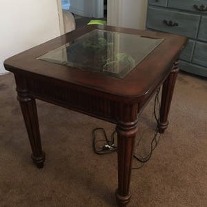 Op de afbeelding: Een donkerbruine houten bijzettafel met een glazen blad. De tafel heeft een gesneden ontwerp op de poten en een kleine plant groeit in het glazen blad.