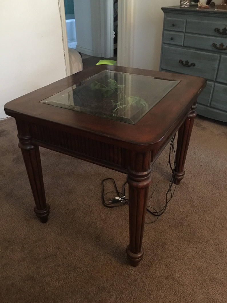 Op de afbeelding: Een donkerbruine houten bijzettafel met een glazen blad. De tafel heeft een gesneden ontwerp op de poten en een kleine plant groeit in het glazen blad.