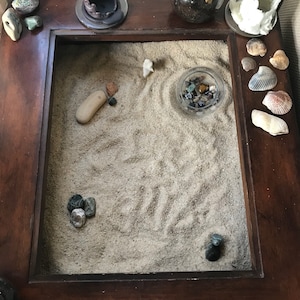 Op de afbeelding: Een houten tafelblad met een zandbak, een kleine kom met gekleurde stenen en verschillende schelpen. De zandbak heeft een klein, wit keramiek figuurtje in het zand.