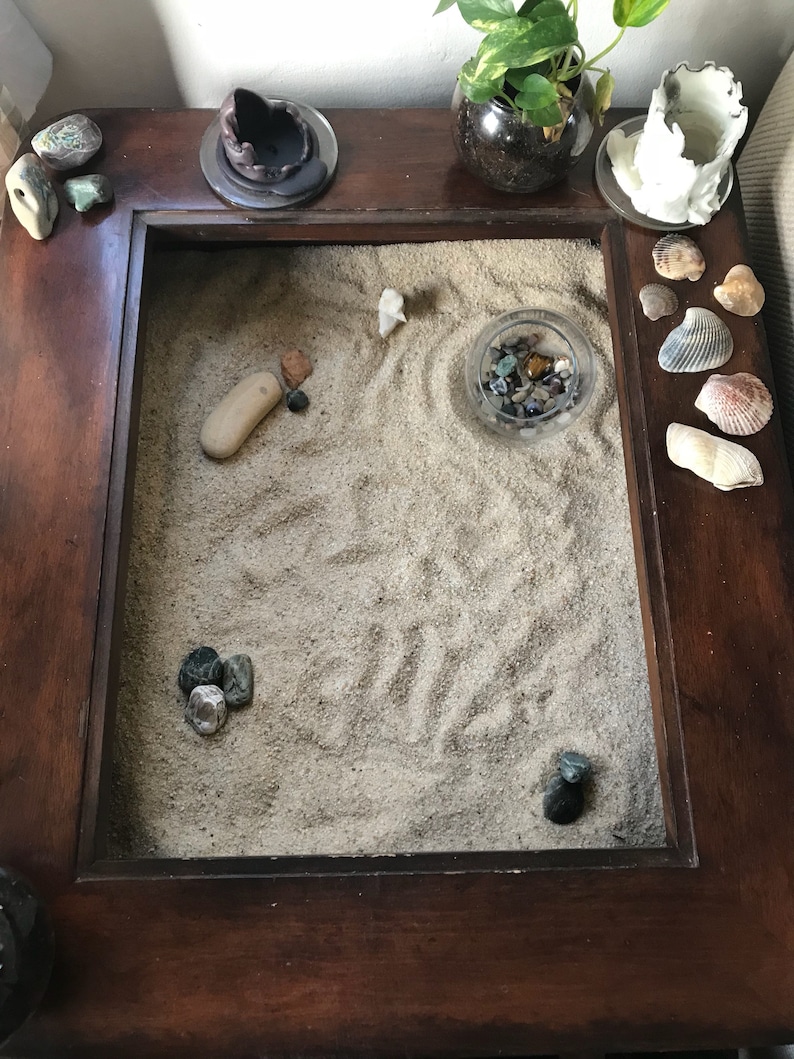 Op de afbeelding: Een houten tafelblad met een zandbak, een kleine kom met gekleurde stenen en verschillende schelpen. De zandbak heeft een klein, wit keramiek figuurtje in het zand.
