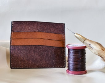 May include: A brown leather card holder with a brown leather thread and a needle. The card holder has multiple slots for cards and is being stitched together.