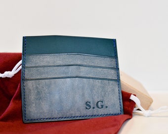 May include: A blue leather card holder with a blue stitching detail and the initials "S.G." embossed on the front.
