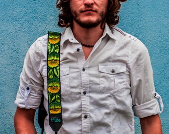 Pode incluir: Um homem vestindo uma camisa branca de botões com uma alça de câmera floral. Ele está em pé na frente de uma parede azul.