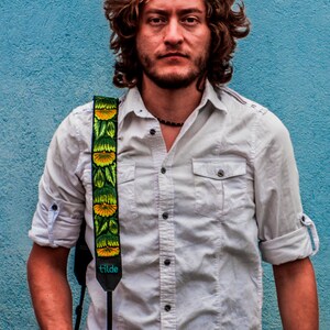 May include: A man wearing a white button-down shirt with a floral camera strap. He is standing in front of a blue wall.