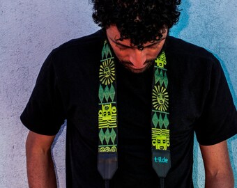 Pode incluir: Uma pessoa vestindo uma camiseta preta com uma alça de câmera estampada em verde e amarelo. A alça tem a palavra "tilde" impressa. A pessoa está segurando uma câmera preta com uma lente.