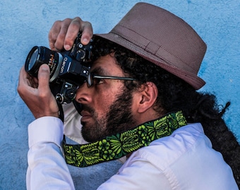 Pode incluir: Uma pessoa usando um fedora marrom e uma camisa branca com uma alça de câmera estampada em verde e preto está segurando uma câmera preta e olhando através do visor.