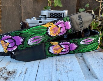 Pode incluir: Uma correia de câmera preta com um padrão floral verde, roxo e amarelo. A correia é bordada com flores e tem um remendo de couro preto com a palavra "tilde" impressa nele.