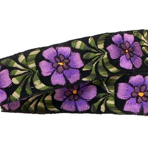 以下が含まれることがあります： 紫色の刺繍の花と緑の葉がついた黒いカメラストラップ。ストラップには、ターコイズ色の刺繍で「tilde」と刺繍された黒い革のパッドが付いています。