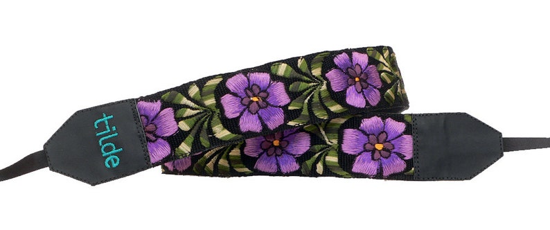 以下が含まれることがあります： 紫色の刺繍の花と緑の葉がついた黒いカメラストラップ。ストラップには、ターコイズ色の刺繍で「tilde」と刺繍された黒い革のパッドが付いています。