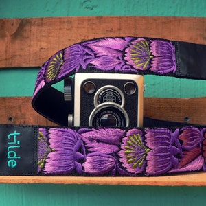 以下が含まれることがあります： 黒革のストラップに「tilde」という刺繍が施された、紫と緑のフローラルカメラストラップです。
