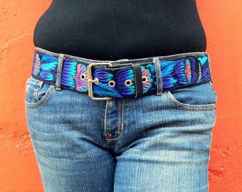 以下が含まれることがあります： 銀色のバックルが付いた青と紫の刺繍のベルト。ベルトは黒いシャツと青いジーンズの上に着用されています。
