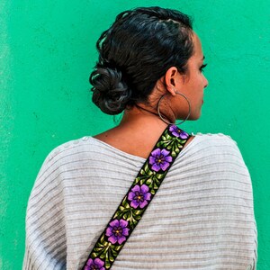 以下が含まれることがあります： グレーのセーターを着た女性が、紫色の花柄の刺繍が施されたカメラストラップをしています。ストラップには「tilde」という文字が印刷されています。
