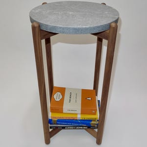 May include: A round, grey, marble-topped side table with a wooden base and four legs. The table is stacked with four books: "The Beach" by Alex Garland, "The Catcher in the Rye" by J.D. Salinger, "The Greek and Roman Myths" by Audrey Hepburn, and "Icons" by Taschen.