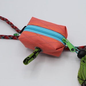 May include: A small coral-colored rectangular pouch with a bright blue zipper and a lime green zipper pull. The pouch is attached to a red and black patterned rope loop and holds a green plastic waste bag that is partially dispensed.