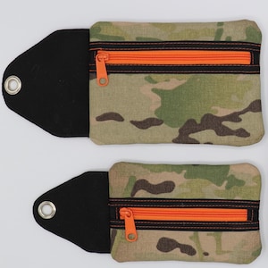 Könnte beinhalten: Eine Sammlung von Pouches und Organisatoren mit Tarnmuster. Die Pouches haben orangefarbene Reißverschlüsse und schwarze Akzente. Die Organizer haben schwarze elastische Bänder und Ösen. Für kleine Gegenstände.