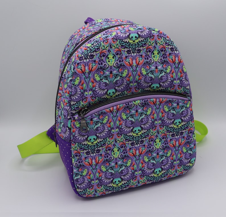 Puede incluir: Mochila morada con un estampado floral caprichoso con detalles verdes y rojos. La mochila tiene dos bolsillos con cremallera en la parte delantera.